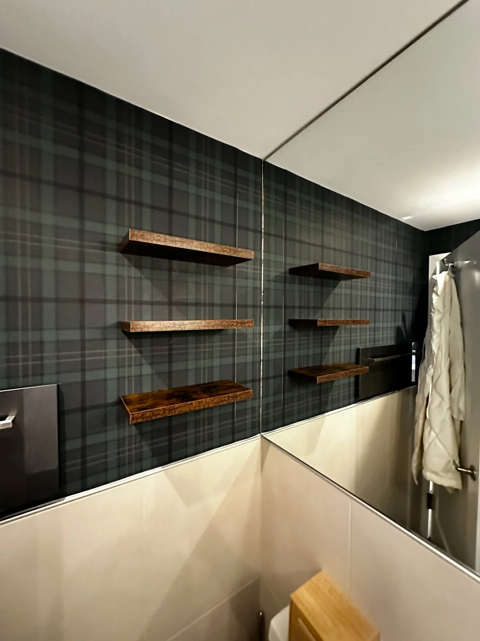 Three wood shelves on a plaid wall beside a mirror reflecting the same shelves